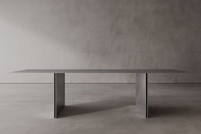 A sleek, modern stainless steel dining table with a minimalist design, featuring a rectangular shape and supported by a central pedestal column.
