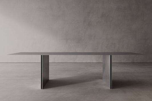 A sleek, modern stainless steel dining table with a minimalist design, featuring a rectangular shape and supported by a central pedestal column.